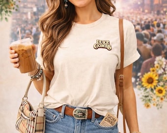 May include: A cream-colored t-shirt featuring the text Luke Combs in brown, paired with distressed blue denim shorts, a brown leather belt with a decorative buckle, a straw cowboy hat, a plaid handbag, and a brown fringe crossbody bag.
