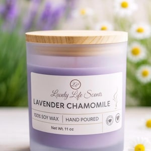 May include: A lavender-colored candle in a frosted glass jar with a wooden lid. The label reads "Lovely Life Scents Lavender Chamomile, 100% Soy Wax, Hand Poured, Net Wt. 11 oz." The background features blurred flowers.