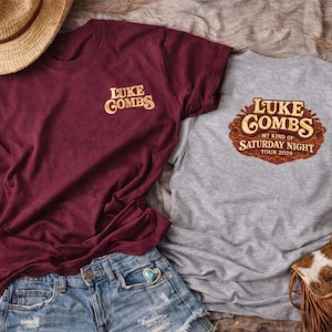 Könnte beinhalten: Zwei T-Shirts mit dem Text "Luke Combs" und "My Kind of Saturday Night Tour 2026". Ein Shirt ist weinrot, das andere grau. Ein Strohhut, Jeansshorts und eine Fransenhandtasche sind ebenfalls zu sehen.