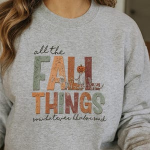 May include: A light grey crewneck sweatshirt featuring the text "all the FALL THINGS" in a distressed, multi-coloured block font. The letter A in FALL is replaced by a small pumpkin illustration. Below the text is the cursive script "sow hatever hhabors oad".