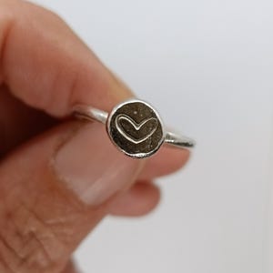 Può includere: Un anello in argento con un elemento centrale circolare grigio scuro testurizzato e un contorno a cuore inciso. La fascia è sottile e lucida, collegata alla montatura arrotondata e leggermente irregolare che mette in risalto il cuore.
