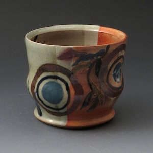 May include: Handmade ceramic cup with a unique design. The cup features a cream-colored base with orange, brown, and blue circular patterns. The rim has orange stripes. The cup is a functional art piece.
