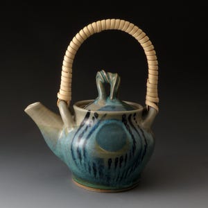 May include: A ceramic teapot featuring a blue and green glaze with dark blue vertical brushstrokes and a circular pattern. The teapot has a curved spout, a lid with a twisted handle, and a tall, arched handle wrapped in light tan woven rattan.
