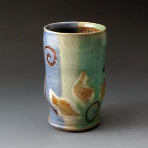 Può includere: Un bicchiere in ceramica fatto a mano di forma cilindrica. Il bicchiere presenta una combinazione di colori blu e verde con disegni a spirale e a forma di foglia marrone. Il bordo interno è di colore marrone scuro.