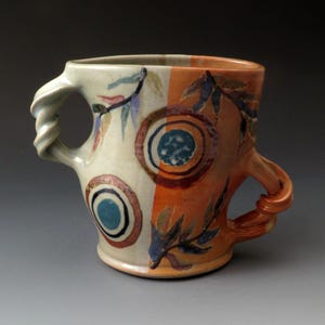 May include: A handcrafted ceramic mug with a distinctive design. The mug is split into cream and orange sections, adorned with circular patterns and leaf motifs. It has two twisted handles. A functional and artistic piece for enjoying hot drinks, made in the UK.