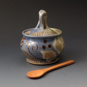 May include: A round ceramic lidded jar with a blue glaze, featuring brown painted swirls and dots, and a looped handle on the lid. The jar has a textured base and is accompanied by a smooth, light brown wooden spoon placed in front of it.