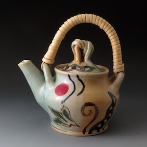 May include: A ceramic teapot with a light beige and pale green glaze, featuring a hand-painted design of a red circle, dark green leaves, and brown swirls. It has a curved spout, a matching lid with a central handle, and a tall, arched rattan handle.
