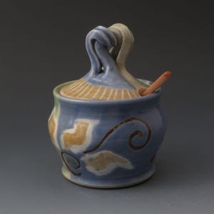 May include: A ceramic sugar bowl with a blue, tan, and white glaze featuring a hand-painted swirl pattern. The lid has a sculpted, twisted handle and a small opening for a light-colored wooden spoon that is resting inside the container.
