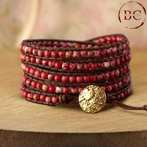 May include: A multi-strand leather wrap bracelet with red and brown jasper beads and a gold heart charm. The bracelet is tied with a leather cord and has a gold clasp.