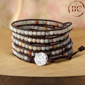 Puede incluir: Un brazalete de envoltura multi-cadena con cordón de cuero marrón y cuentas de color verde claro, azul y marrón. El brazalete tiene un dije de plata con un diseño de hoja.
