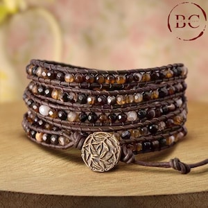 May include: A brown leather wrap bracelet with multiple strands of brown, black, and white agate beads. The bracelet has a decorative metal clasp with a leaf design.