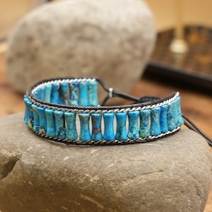 Op de afbeelding: Een blauwe en zilveren kralen armband met een zwart koord en een verstelbare sluiting. De armband is gemaakt van kleine, cilindrische kralen met een marmerpatroon.