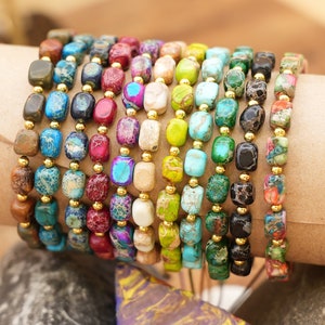May include: A collection of beaded bracelets featuring a variety of colorful, rectangular stones. The stones are in shades of blue, green, red, and brown, strung together with gold-colored beads. The bracelets are stacked together, showcasing the different color combinations.