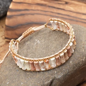 May include: A beige and white beaded bracelet with a gold-coloured cord. The bracelet is made of small, rectangular beads with a marbled pattern.