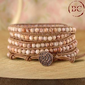 Puede incluir: Un brazalete de cuero de múltiples vueltas con un cierre de cobre. El brazalete presenta múltiples hileras de cuentas de color marrón claro y blanco.