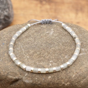 May include: A gray and silver beaded bracelet with a gray cord closure. The bracelet is made of small, round beads with silver accents.