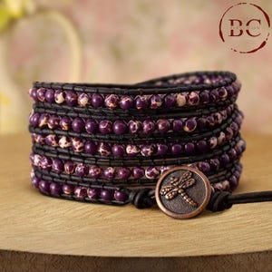 May include: A multi-wrap bracelet with purple and white beads and a dragonfly charm. The bracelet is made of leather and has a brown clasp.