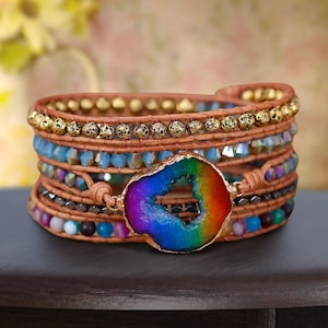 May include: Multi-layered bracelet with a rainbow agate charm. The bracelet features brown leather cord, gold beads, blue crystals, and black beads.