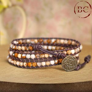 May include: Wrap bracelet featuring a combination of orange, white, and brown beads. The bracelet is made with multiple strands of beads and leather, secured with a button clasp. The bracelet is designed to wrap around the wrist multiple times.