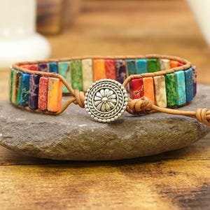 May include: A multi-colored leather bracelet with a silver flower charm. The bracelet is made with rainbow-colored stones and has a brown leather strap.
