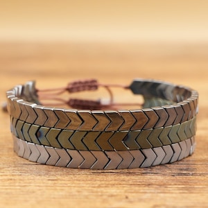 May include: A layered bracelet with a chevron design. The bracelet features three rows of metallic, arrow-shaped beads in silver and bronze tones. The adjustable cord is a light brown color.