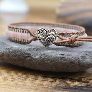 May include: A wrap bracelet with pink beads and brown leather straps. The bracelet features a silver heart-shaped clasp with an ornate design. The bracelet is displayed on a gray stone surface.