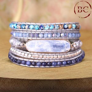 Puede incluir: Un brazalete de varias capas con cuentas azules y plateadas, una piedra rectangular azul claro y un dije plateado con las letras "BC" en un círculo.
