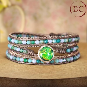 Könnte beinhalten: Ein mehrreihiges Wickelarmband mit grünen und blauen Perlen und einer goldfarbenen Kette. Das Armband hat einen runden, grün-goldenen Opal-Stein in der Mitte.