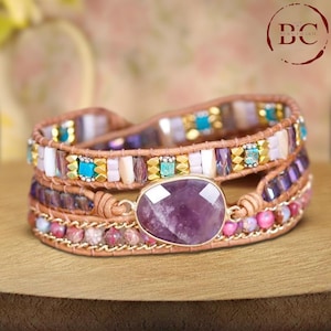 Puede incluir: Un brazalete de envoltura de varias capas con un gran amuleto de amatista. El brazalete está hecho de cuero marrón, cuentas doradas y piedras de varios colores.