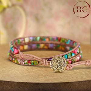 May include: A wrap bracelet with colorful square beads in shades of green, pink, purple, and orange. The bracelet is secured with a light brown leather cord and a gold-colored button clasp with a leaf design. The bracelet is on a wooden surface.