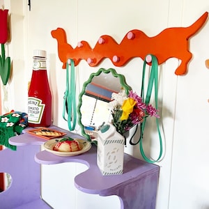 May include: A purple wooden bench with two shelves. The top shelf has a plate with a red dessert and a ketchup bottle labeled "Heinz Tomato Ketchup". The bottom shelf has a white milk carton labeled "Original". A bright orange wooden dachshund-shaped coat rack with pink knobs is mounted on the wall behind the bench. The rack has two green leashes hanging from it. A mirror with a scalloped edge is on the wall behind the bench. A bouquet of yellow and pink flowers is in front of the mirror.