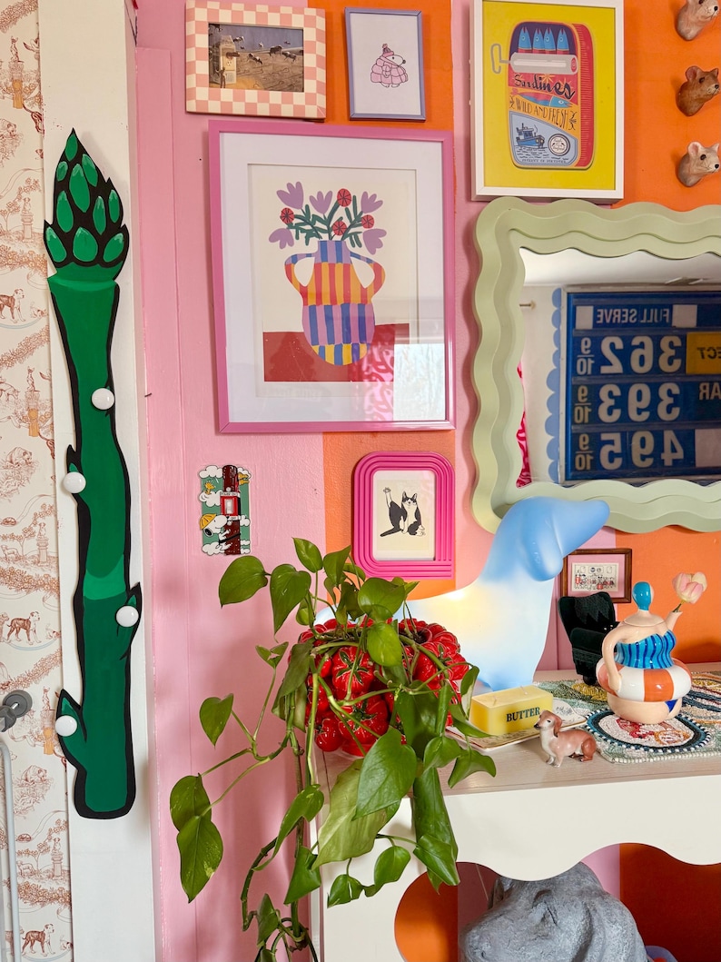 Op de afbeelding: Een groene asperge-vormige wanddecoratie met witte stippen, een roze muur met een ingelijst plaatje van een gestreepte vaas met bloemen, een wit keramiek hondenbeeldje, een plant in een rode pot en een witte tafel met een botervloot en een klein hondenbeeldje.