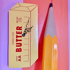 May include: A yellow stick of butter clock with a black clock face and hands. The butter stick is labeled "Grade AA Butter Unsalted Net Wt. 4 oz (113g)".