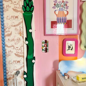 Op de afbeelding: Een groene asperge-vormige wandhaak met witte ronde knoppen. De haak is gemonteerd op een roze muur met een wit en bruin gestreept behang. Er zijn andere decoratieve items op de muur, waaronder een ingelijst afdruk van een vaas met bloemen en een ingelijst afdruk van een kat.