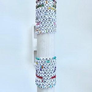 Puede incluir: Una colecci&oacute;n de pulseras de cuentas con letras blancas que deletrean varias palabras y frases. Las pulseras de colores se exhiben en un soporte cil&iacute;ndrico blanco.