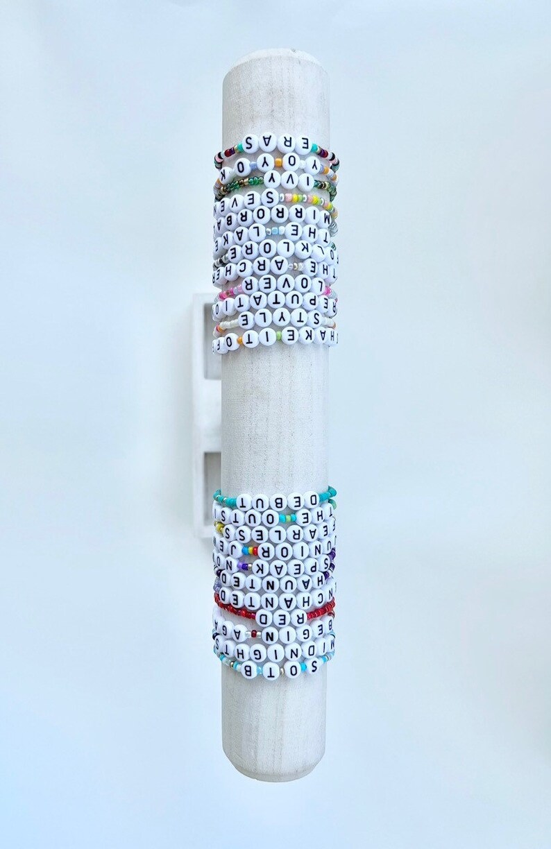 Puede incluir: Una colecci&oacute;n de pulseras de cuentas con letras blancas que deletrean varias palabras y frases. Las pulseras de colores se exhiben en un soporte cil&iacute;ndrico blanco.