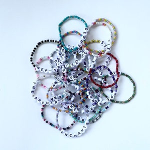 Puede incluir: Una colecci&oacute;n de pulseras de cuentas de colores, algunas con cuentas de letras blancas que deletrean palabras como "THE ARCHER" y "ENCHANT". Las pulseras est&aacute;n hechas de peque&ntilde;as cuentas de colores.