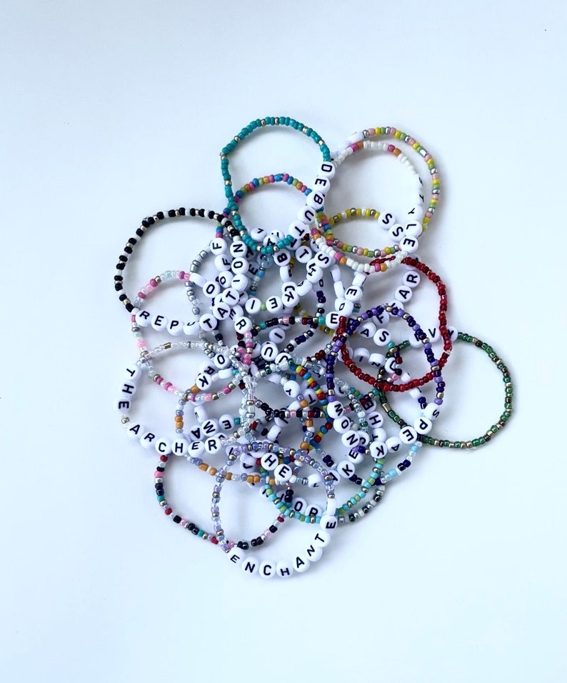 Puede incluir: Una colecci&oacute;n de pulseras de cuentas de colores, algunas con cuentas de letras blancas que deletrean palabras como "THE ARCHER" y "ENCHANT". Las pulseras est&aacute;n hechas de peque&ntilde;as cuentas de colores.