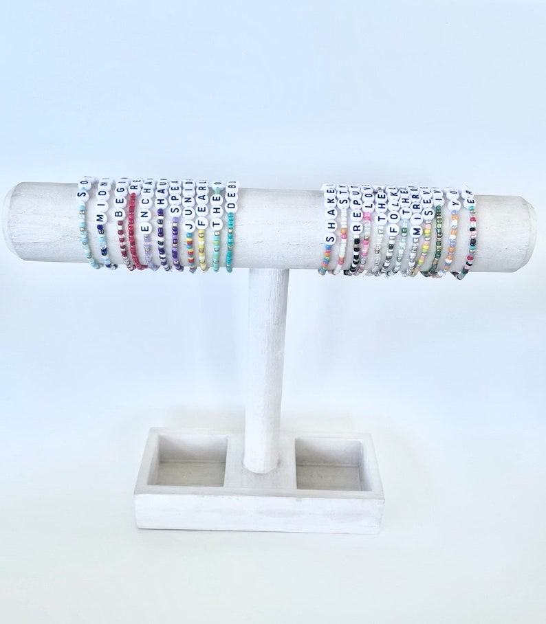 Puede incluir: Una exhibici&oacute;n de pulseras de cuentas con cuentas de letras blancas que deletrean palabras como "Midnight", "Enchanted" y "Shake". Las pulseras son de varios colores y se exhiben en un soporte de madera blanco.