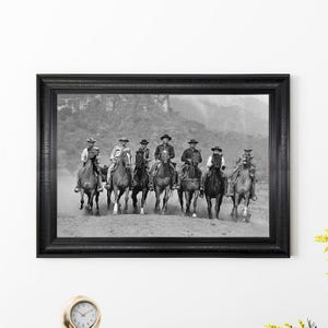 以下が含まれることがあります： 白黒の額入りプリント。カウボーイのグループが馬に乗っています。カウボーイは帽子をかぶり、埃っぽい風景の中を馬に乗っています。プリントは黒いフレームに入っています。