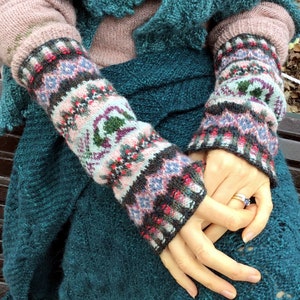 Könnte beinhalten: Ein Paar gestrickte Armwärmer mit einem farbenfrohen geometrischen Muster in den Farben Rosa, Blau, Grün und Schwarz. Die Armwärmer werden über einem rosa Pullover und einem türkisfarbenen Strick-Schal getragen.