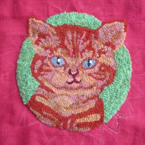 May include: A close-up of a red, orange, and green needlepoint cat with green eyes. The cat is stitched on a red fabric background.