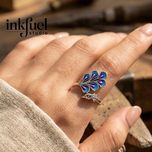 May include: A silver-toned ring featuring a leaf design with blue and purple enamel detailing. The ring has a bird-like element at the base. The image includes the text "inkfuel studio".