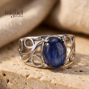 Puede incluir: Un anillo de plata con una piedra preciosa ovalada de color azul oscuro. El anillo tiene un diseño calado con patrones curvos y circulares. La piedra preciosa es el punto focal, engastada en una banda de plata pulida. El anillo se muestra sobre una superficie texturizada de color marrón claro.