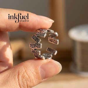 May include: A silver-colored ring with a repeating rectangular design. Each rectangle features a swirling pattern. The ring is held between fingers, with the "inkfuel studio" logo visible in the background.