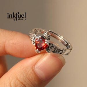 May include: A silver ring featuring a heart-shaped, red gemstone. The ring has a crown and wing design, with the text "inkfuel studio" visible. The ring is adjustable and has the number "925" engraved on it.