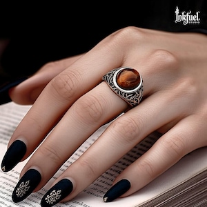 Puede incluir: Un anillo de plata con una piedra preciosa ovalada y marrón. El anillo tiene un diseño de filigrana ornamental alrededor de la piedra. El logotipo de Inkfuel Studio es visible. La mano tiene esmalte de uñas negro con detalles dorados.