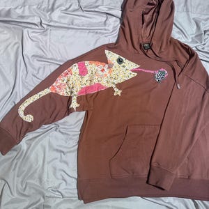May include: Brown hooded sweatshirt with a large chameleon applique on the front and left sleeve. The chameleon is made of floral and pink patterned fabric. The sweatshirt has a front pocket and a drawstring hood.