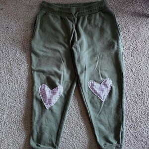Puede incluir: Pantalones deportivos verde oliva con dos parches en forma de corazón en las rodillas. Los parches son de tela a cuadros blanca y morada. Los pantalones tienen cintura con cordón y bolsillos.