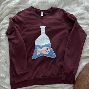 May include: A burgundy sweatshirt featuring an applique of a fish in a bottle. The fish is peach-coloured with a smiling face, set against a blue quilted background. The bottle is white. A casual, comfortable top.
